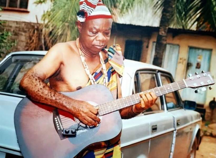 Elderly Mike Ejeagha sitting down shirtless with a guitar. Credit: von.gov.ng